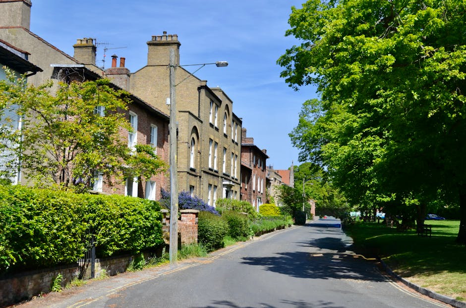 A quiet residential street during daytime with a clear blue sky, lined with multi-storey brick houses featuring large windows and chimneys. The houses are surrounded by well-maintained front gardens w